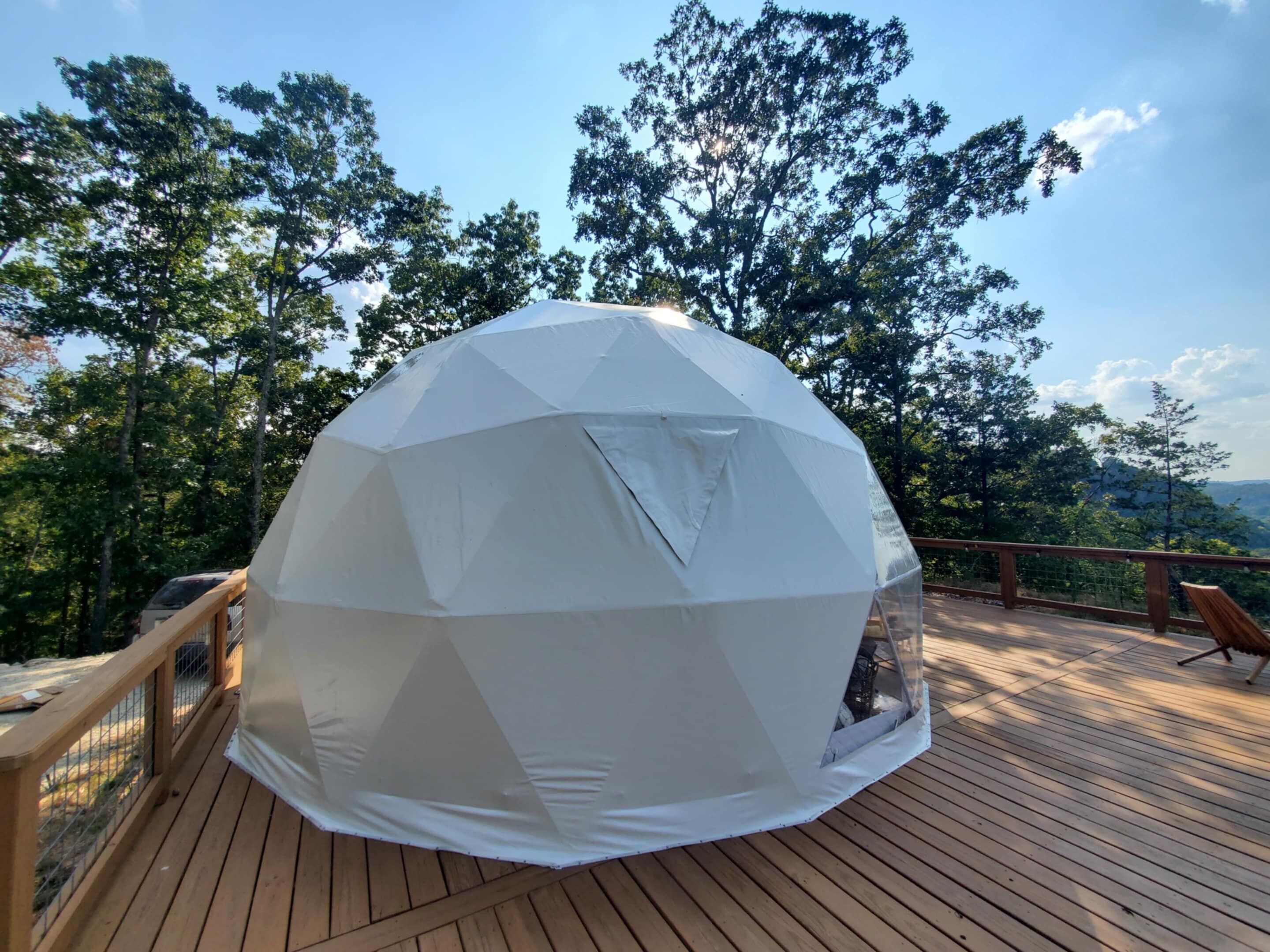 Une structure en forme de dôme géodésique, construite sur une terrasse en bois dotée d’une rambarde, entourée d’arbres et surplombant un ciel dégagé.