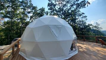 Une structure en forme de dôme géodésique, construite sur une terrasse en bois dotée d’une rambarde, entourée d’arbres et surplombant un ciel dégagé.