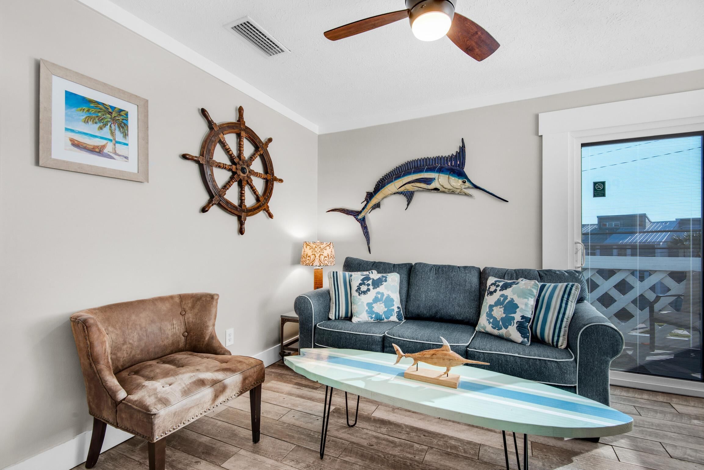 A living room with a brown leather armchair, a blue sofa with patterned cushions, a wooden ship wheel, a large fish mural, and a surfboard on a glass table.