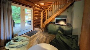 A log cabin interior with a wooden staircase, a television on a cabinet, a sofa, and a sliding glass door leading to an outdoor area. - The Loghouse between Ghent and Bruges (Aalter)