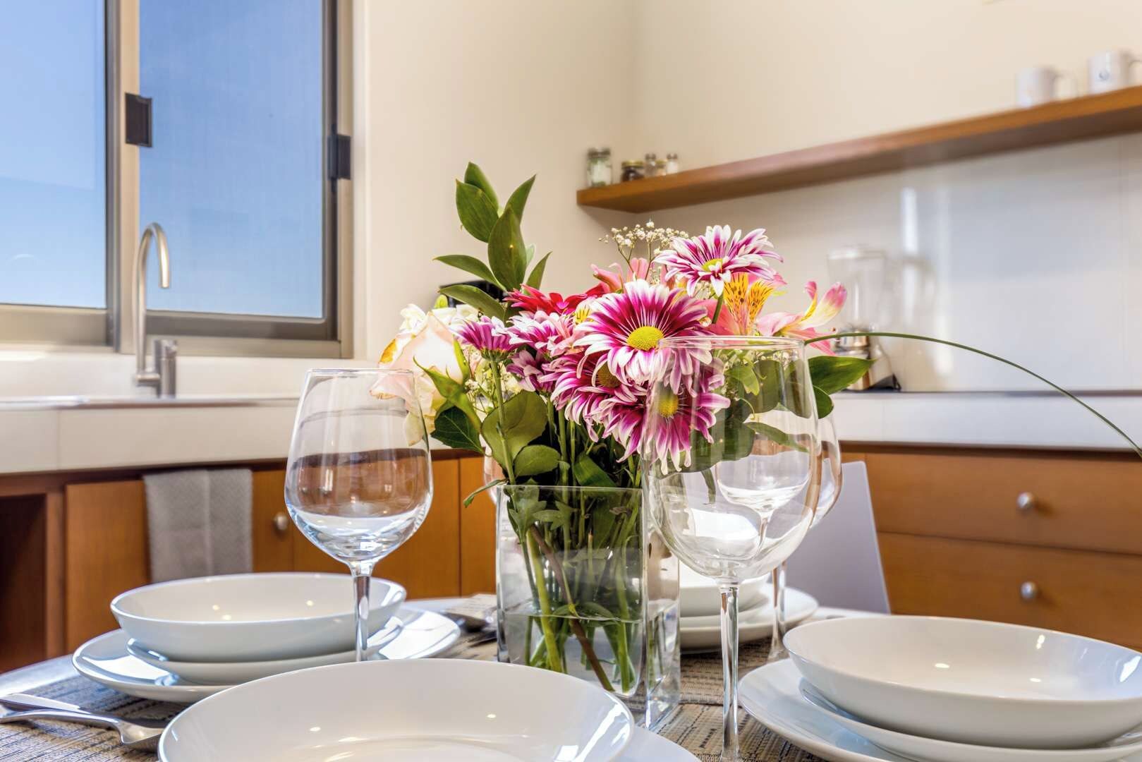 Une table à manger dressée avec de l’assiette blanche, deux verres à vin et un vase contenant des fleurs roses et blanches.