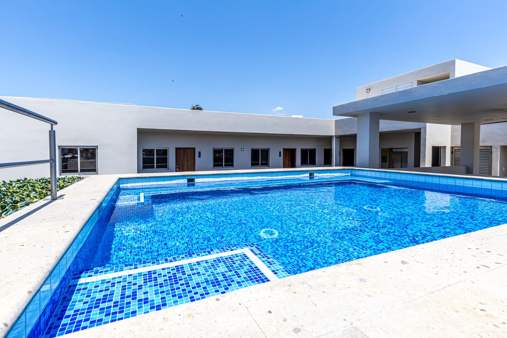 A rectangular swimming pool with blue tiles, surrounded by a white building with multiple windows and doors.
