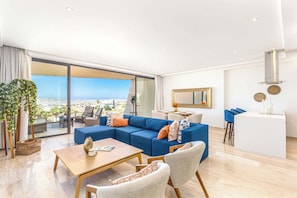 A modern living room with a blue sectional sofa, a wooden coffee table, and a dining area with blue chairs. - Luxury 2 BR Condo - Oceanview Pool - Gym (El Tezal)