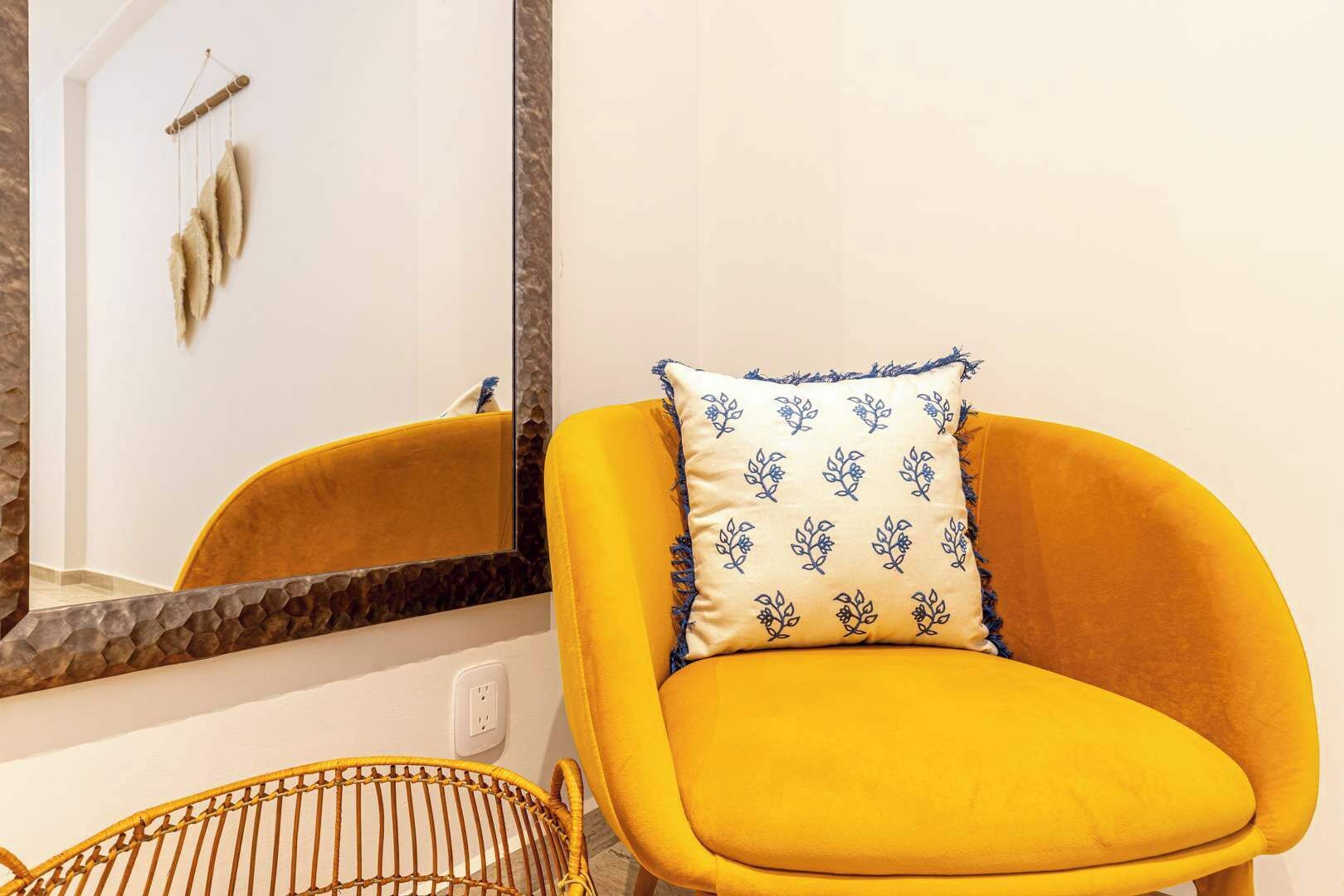 Een gele fauteuil met een gekleurd kussen voor een spiegel met houten lijst.