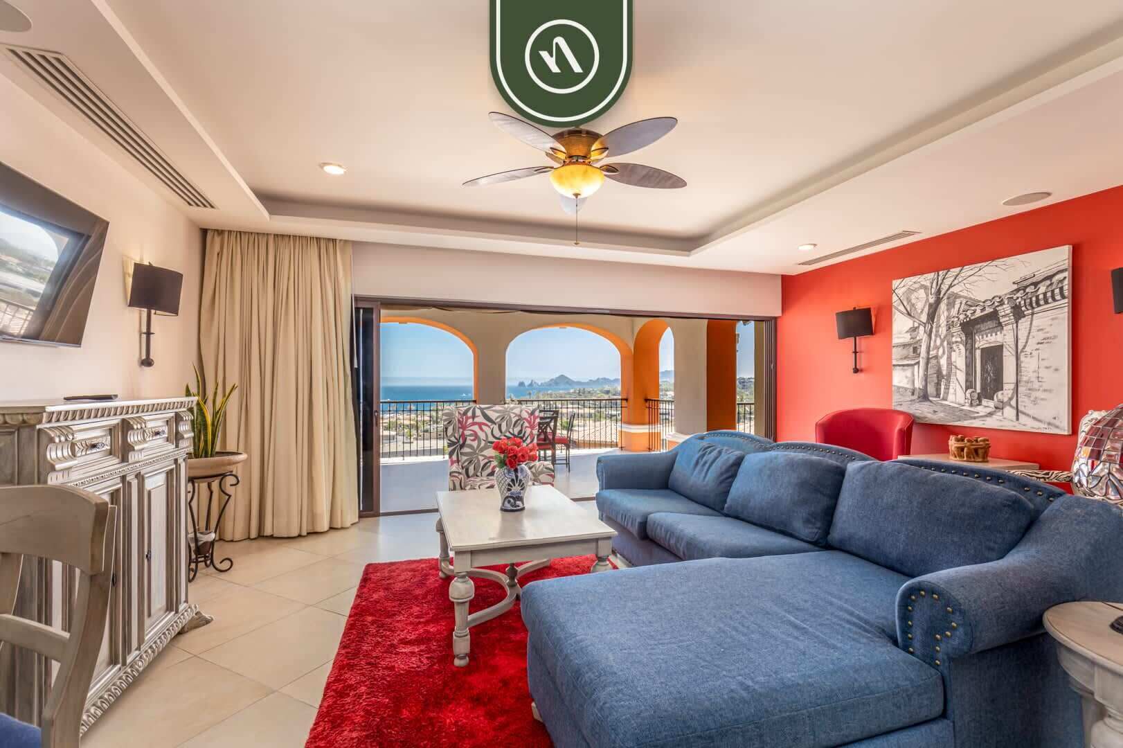 A modern living room with a blue sofa, a red rug, and a balcony with a view of the sea.