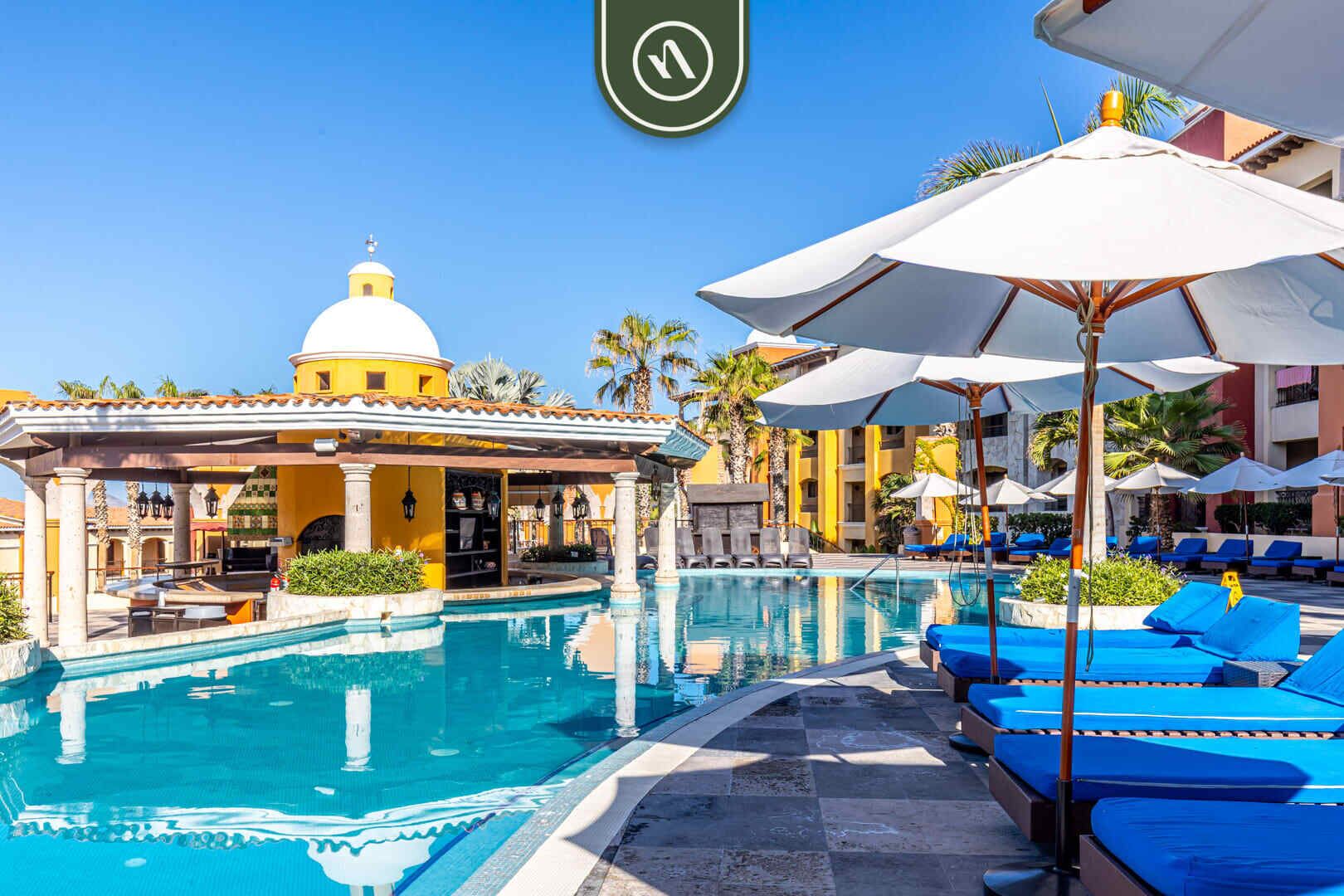 A pool area with blue lounge chairs and umbrellas, a building with a dome, and a clear blue sky.