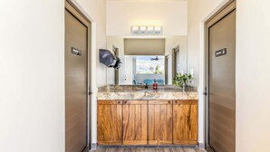 A bathroom with a wooden vanity, a mirror, a ceiling fan, and a view of the city through the window. - Centric Apt in Downtown - Rooftop Pool - Terrace (Cabo San Lucas)