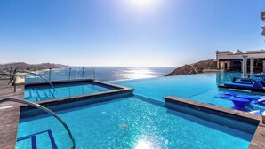 A pool with a view of the ocean, a lounge area with blue chairs, and a building with columns.