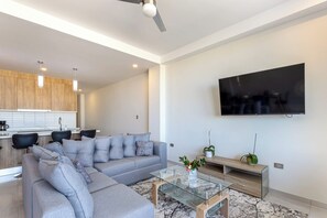 Una sala moderna con un sofá seccional, una mesa de centro de vidrio, un televisor de pantalla plana montado en la pared y una cocina al fondo.