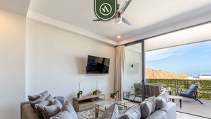 A modern living room with a grey sofa, a flat-screen TV, and a glass coffee table. There is a balcony with outdoor seating and a view of the landscape. - Splendid 2 BR Condo with Private Terrace - BBQ (Cabo San Lucas)