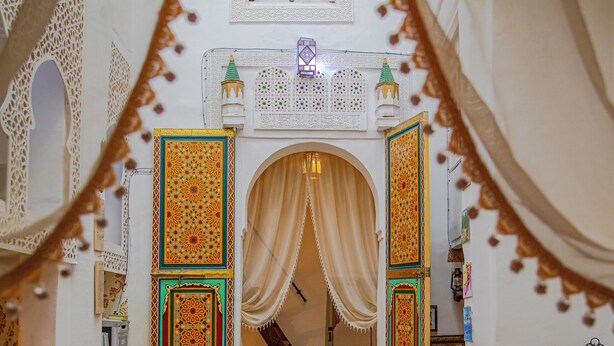 An interior with a dining area featuring tables and chairs, a staircase, and ornate doors with intricate designs. - RIAD MOHAMADIA (Fes)