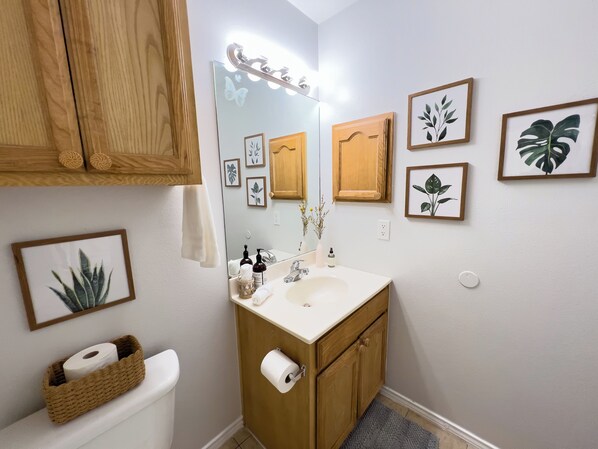 A bathroom with a wooden vanity, a mirror with lights, framed plant pictures, and a toilet with a basket of toilet paper.