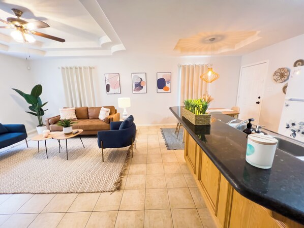 Una sala de estar moderna con un sofá marrón, sillones azules, una mesa de centro y una planta grande. Se observa también una cocina con encimera negra y un fregadero.