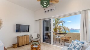 A modern living room with a flat-screen TV, wooden furniture, a balcony with a view of the sea, and a ceiling fan.
