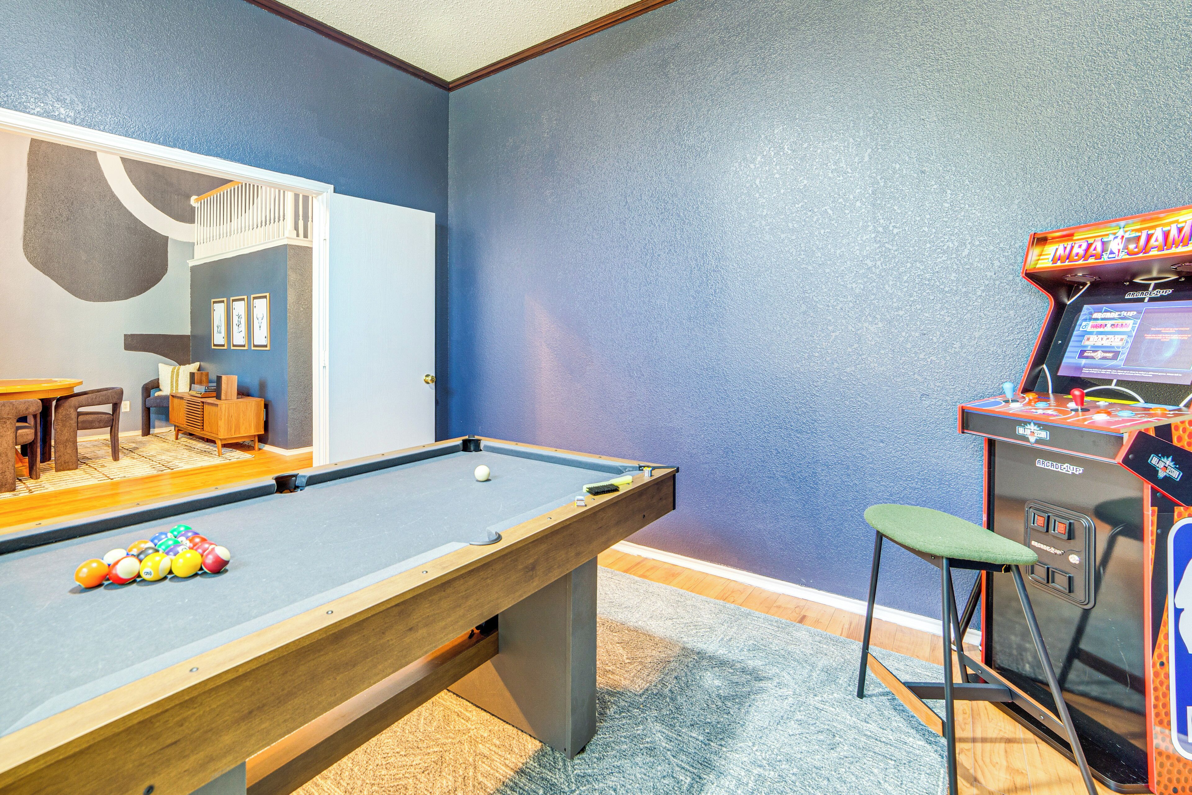Une table de billard avec des billes, un petit tabouret et une autre table de billard dans une pièce aux murs bleus et au plancher en bois.