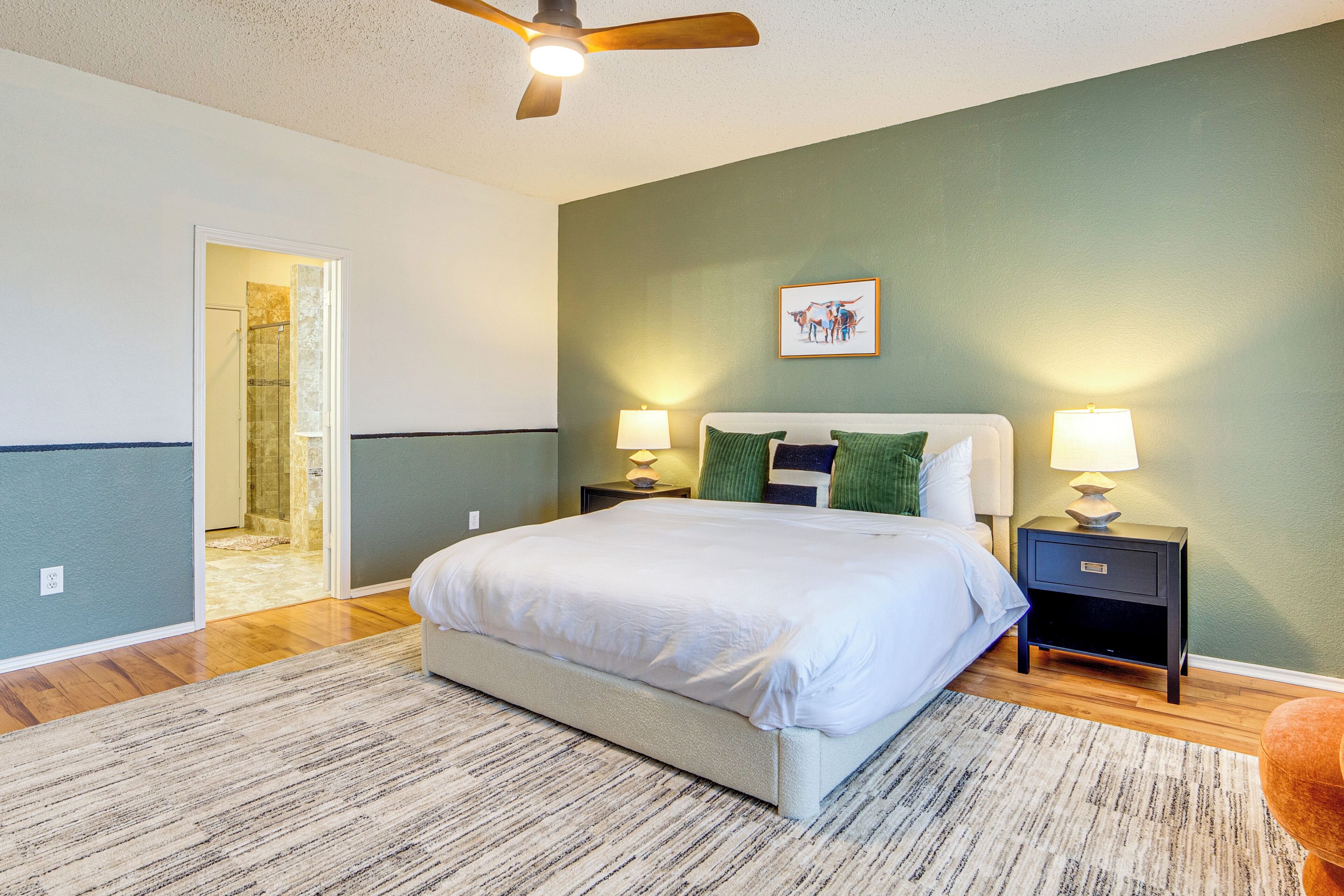 A bedroom with a bed, two bedside tables with lamps, a ceiling fan, and a door leading to another room.