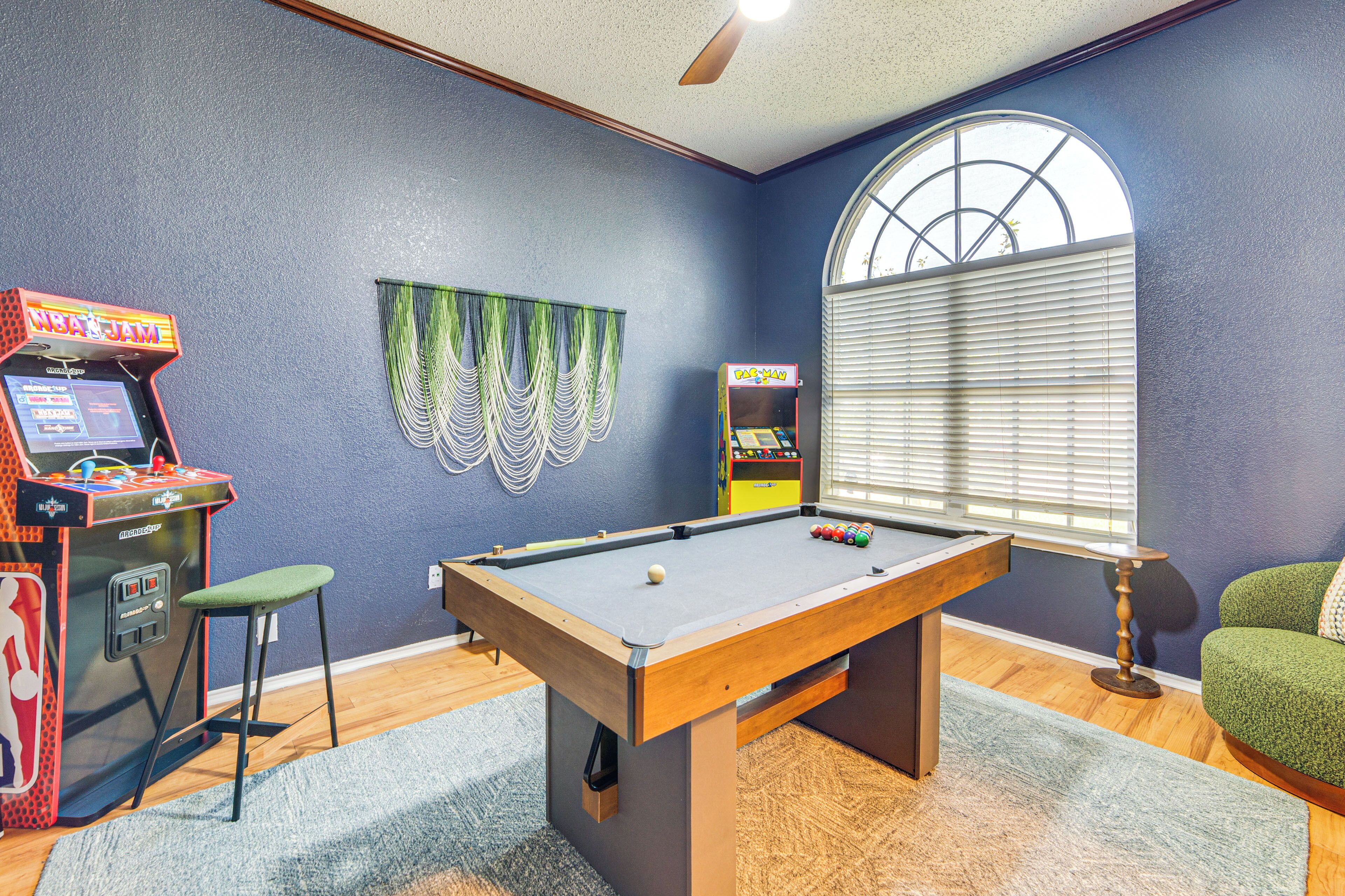 A room with a pool table, arcade games, and a window with blinds.