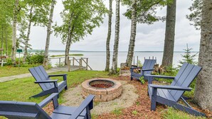 Un espace extérieur aménagé avec des chaises Adirondack et un foyer, entouré d’arbres et offrant une vue sur un plan d’eau.