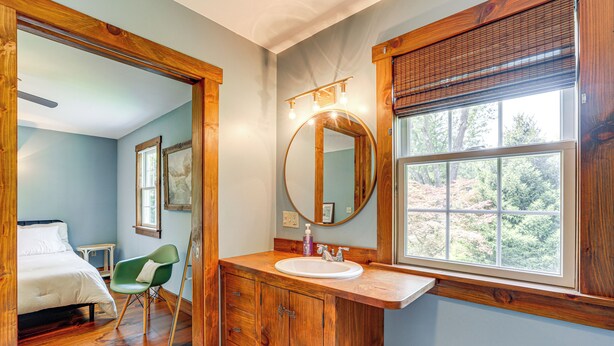 Une chambre à coucher avec un lit, un fauteuil vert, une coiffeuse en bois avec un miroir et une fenêtre donnant sur des arbres.