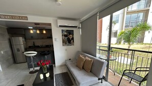 Ein modernes Apartment mit grauem Sofa, einem Esstisch mit roten Blumen, einer Küche und einem Balkon mit Blick auf ein Gebäude und Grünflächen.