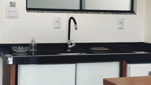 A modern kitchen with a black countertop, a stainless steel sink, and a mirror above the sink. There are two electrical outlets on the wall, and a small dish rack on the countertop. - Pousada Rosa Paradise (Imbituba)