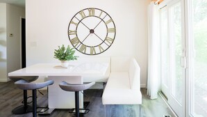 Un comedor moderno con una mesa blanca, dos taburetes de bar, una planta en maceta y un reloj de pared grande.