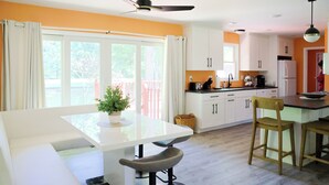 Una cocina moderna con armarios blancos, un comedor con una mesa blanca y dos taburetes de bar, y un ventanal con cortinas.