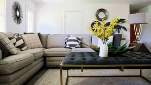 Una sala de estar con un sofá modular, un taburete tapizado, un jarrón con flores amarillas y una lámpara de pie moderna.