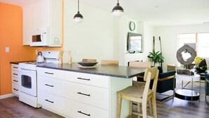 Una cocina moderna con armarios blancos, un microondas integrado y una isla grande con un recipiente. Hay una silla de madera y una mesa pequeña con una planta.