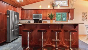A kitchen with wooden cabinets, a stainless steel refrigerator, a microwave, a sink, and a bar area with stools. - Cozy 3BR Lakefront Retreat Featuring Sauna, Hot Tub, Kayaks (Ditch Lake)