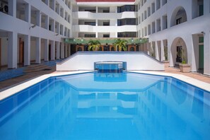 A swimming pool with a central diving area, surrounded by a multi-level building with balconies and potted plants.