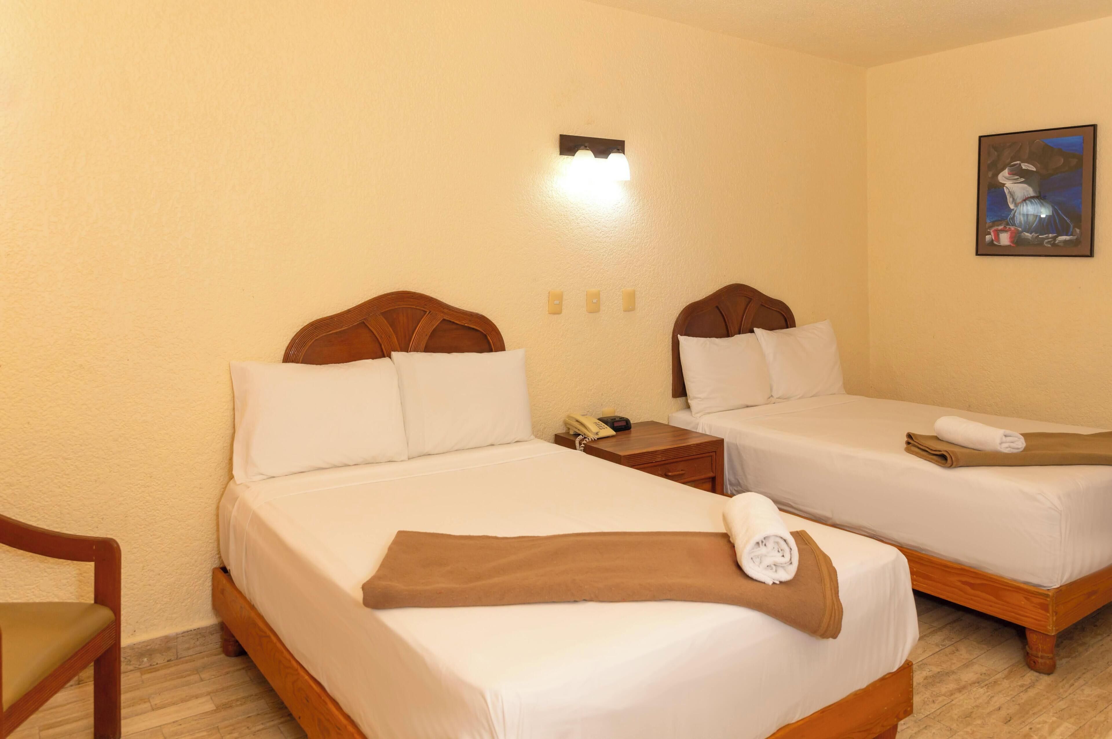 Quarto de hotel com duas camas, cabeceira de madeira, mesa de cabeceira, uma cadeira e um quadro na parede.