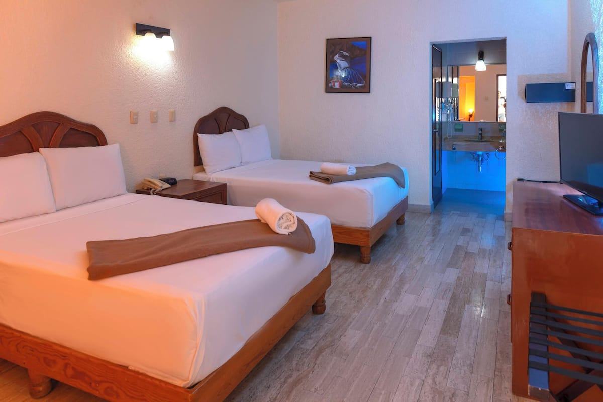 Habitación de hotel con dos camas, un escritorio, un televisor y un baño visible a través de una puerta abierta.
