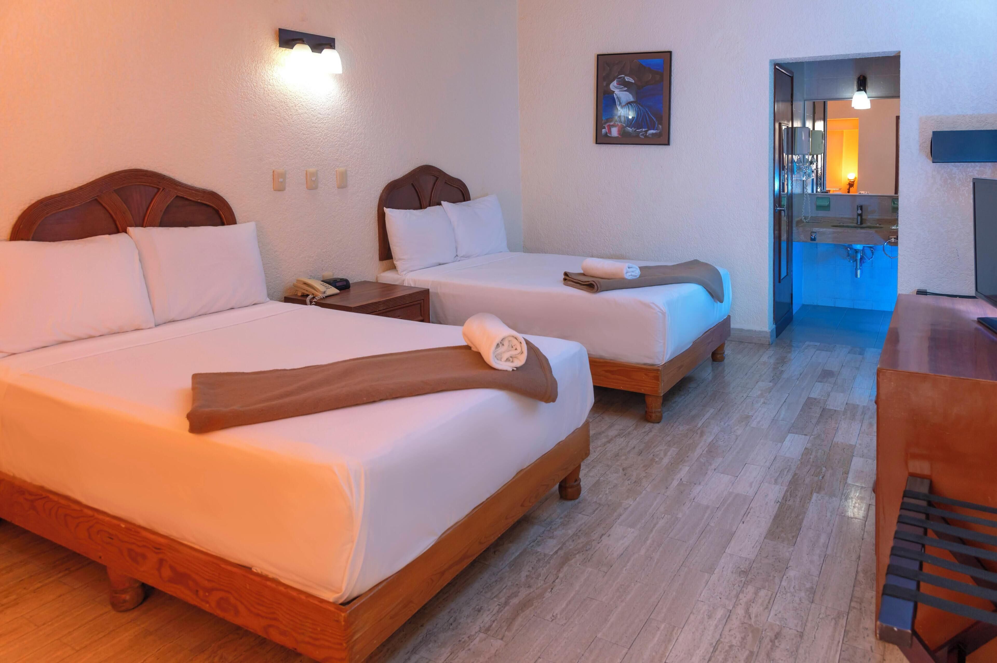 A hotel room with two beds, a desk, a chair, a nightstand, a painting, and a bathroom visible through an open door.