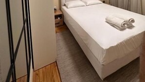A neatly made bed with white linens and a rolled towel, a bedside table with a lamp, and a ceiling light. - Check Point - Acropolis Temple (Athens)