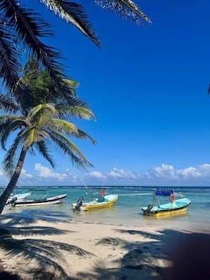 Una playa con tres barcos anclados cerca de la orilla, palmeras y un cielo azul despejado.