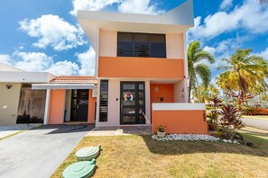 A modern two-story building with a flat roof, a black door, and a small garden with palm trees and a white wall.