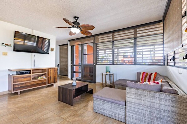 A modern living room with a flat-screen TV, wooden entertainment unit, a ceiling fan, and a large window with blinds.