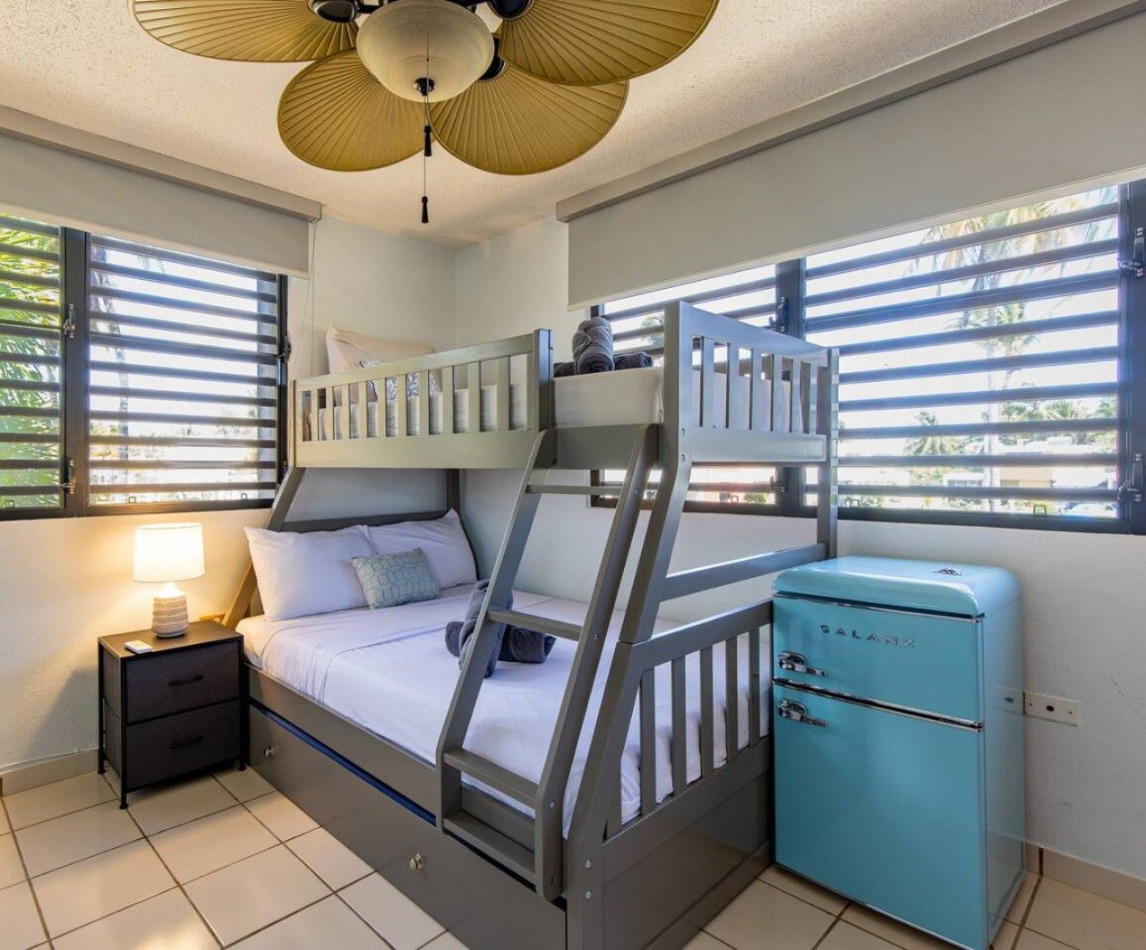 Quarto com beliche, um ventilador no teto, uma mesinha com abajur e um eletrodoméstico azul.
