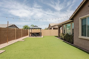 A backyard with artificial grass, a covered patio area, a brick wall, and string lights.