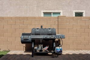 Una barbacoa portàtil amb una ampolla de gas propà i una bossa de carbó al davant d'una paret de maó.
