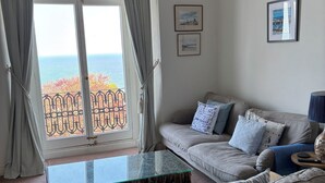 A living room with a sofa, a glass coffee table, and a view of the sea through a window with curtains. - Hillsborough Terrace (Ilfracombe)