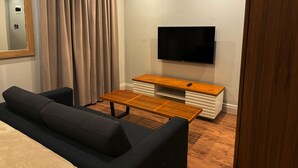 Un salon moderne avec un canapé, une table basse, un téléviseur posé sur un meuble en bois et une grande fenêtre avec des rideaux.