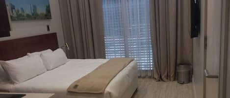 A hotel room with a bed, a bedside table, a TV mounted on the wall, and a window with curtains.