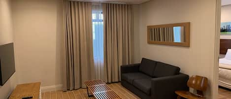 A modern hotel room with a dark grey sofa, a wooden coffee table, a wooden chair, a flat-screen TV mounted on the wall, and a window with beige curtains.