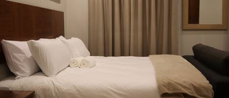 A neatly made bed with white linens and a beige throw blanket, a wooden nightstand with a drawer, and a dark-colored sofa.