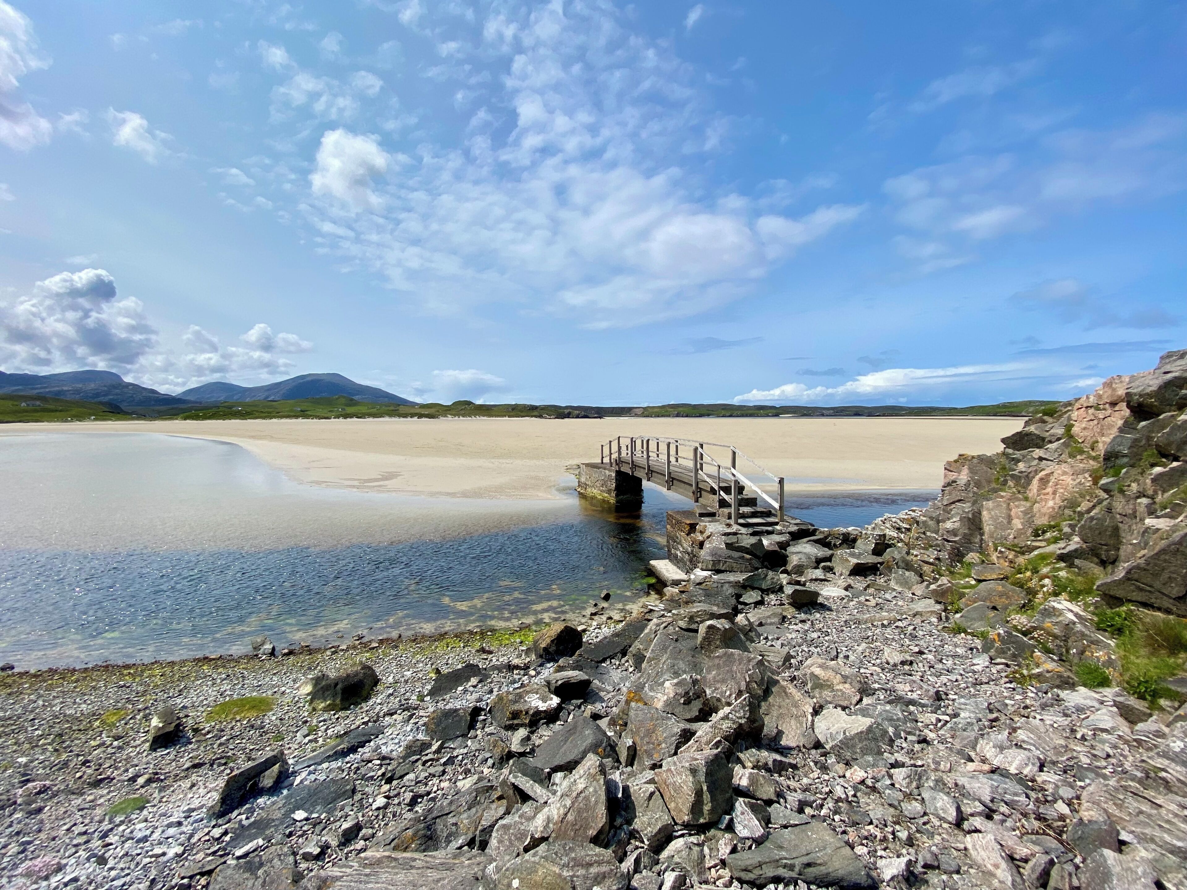 A rocky shoreline with a wooden bridge leading to a sandy beach and mountains in the background.