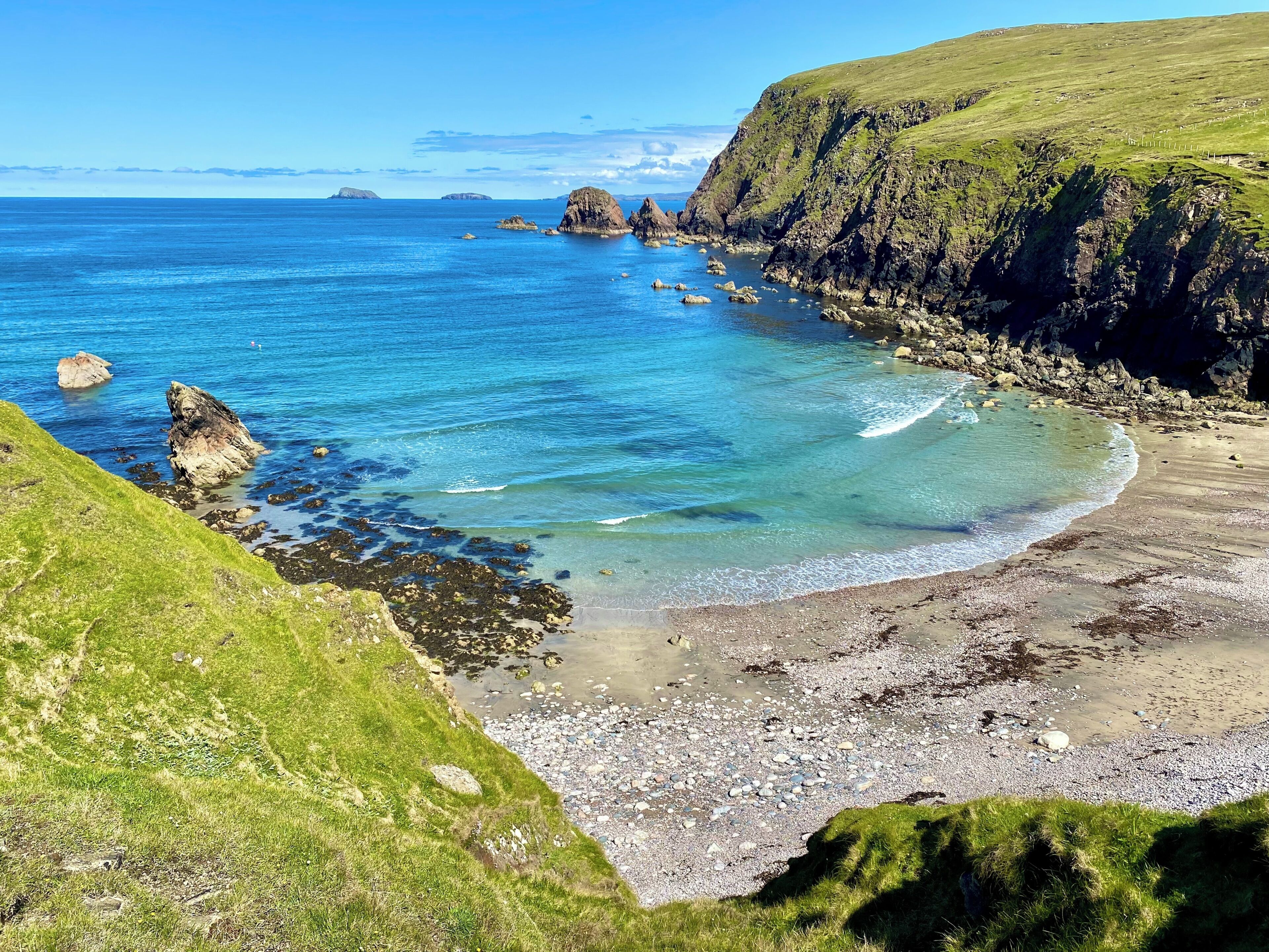 A coastal landscape with clear blue waters, rocky shorelines, and grassy hills.