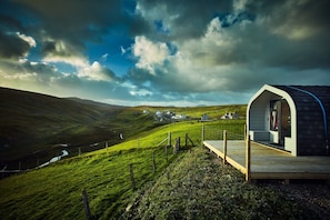 A modern, single-story cabin with a wooden deck and a large window, set in a lush green valley with a view of distant houses and a cloudy sky.