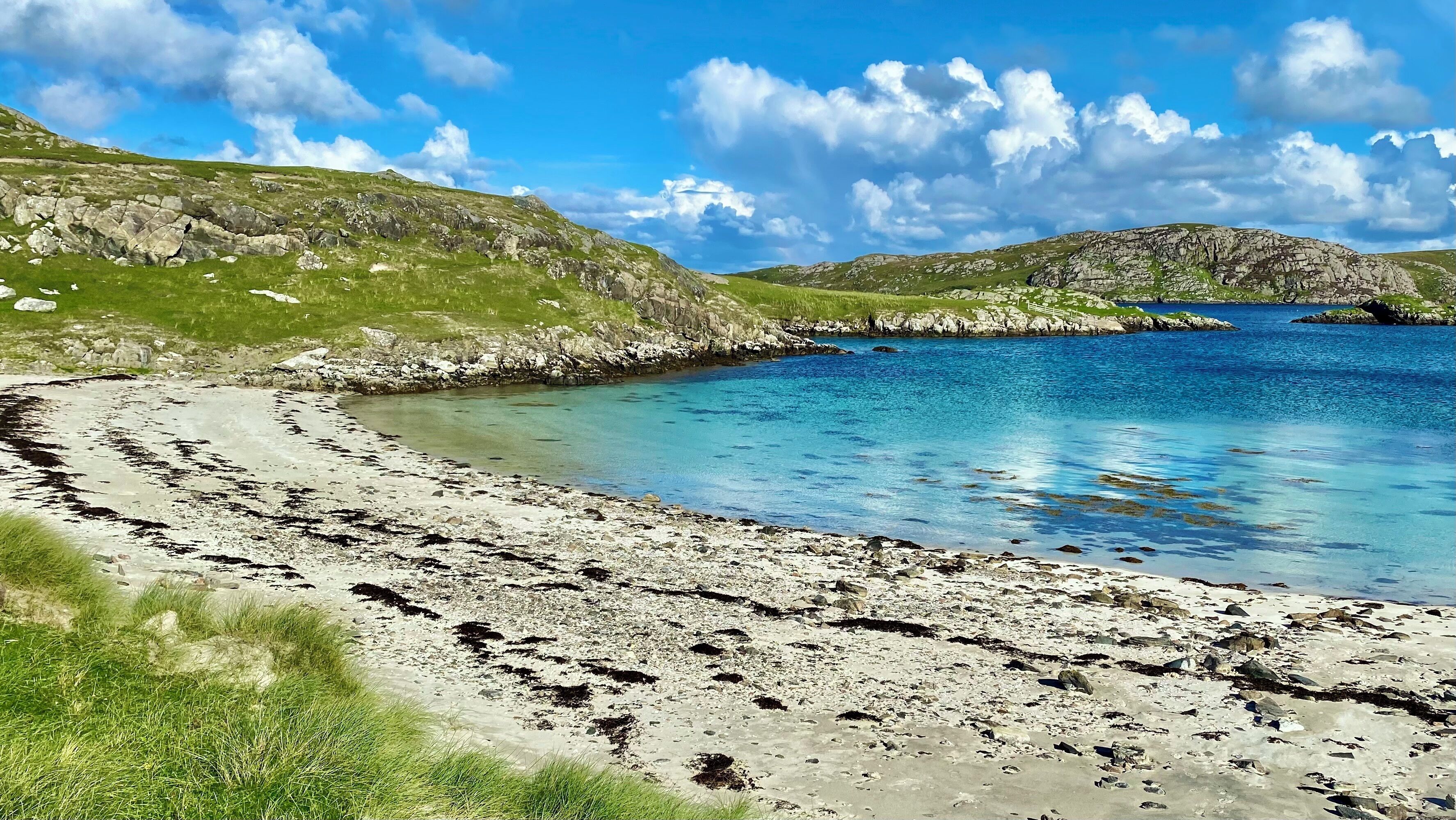 A coastal landscape with a clear sky, rocky terrain, and a sandy beach leading to calm waters.